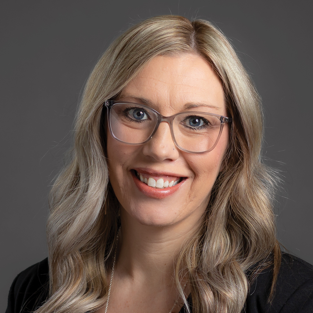 Smiling woman with long wavy blonde hair and glasses, wearing a black blazer against a dark gray background.