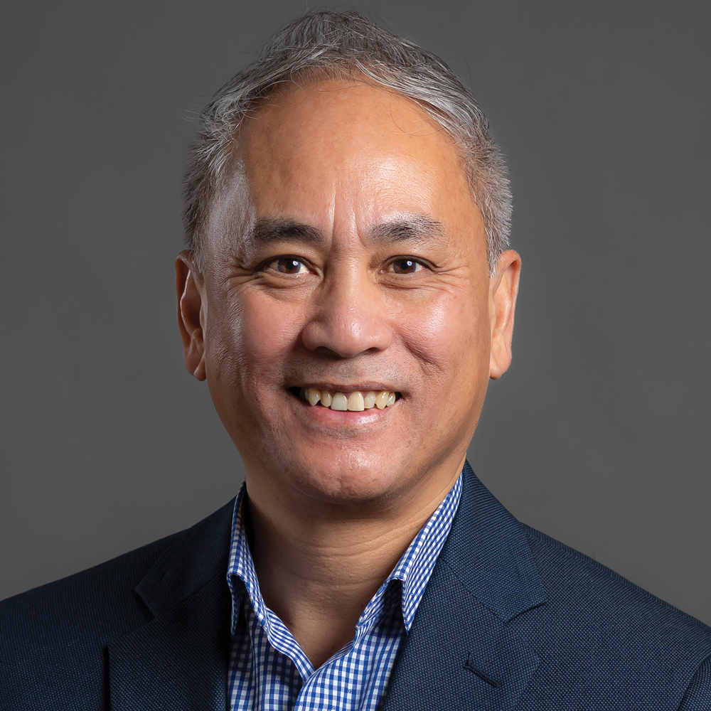 Smiling man with short salt-and-pepper hair, wearing a navy blazer and blue-and-white checkered shirt against a dark gray background.