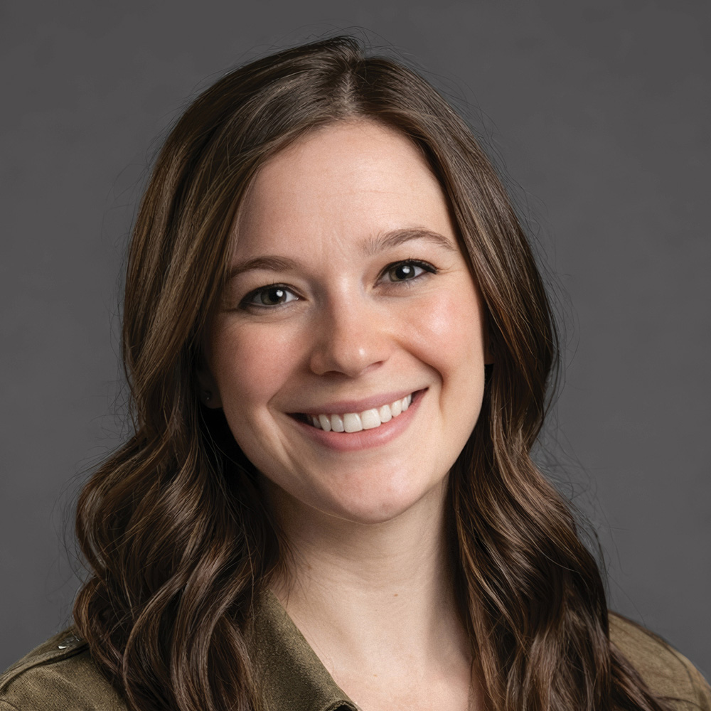 Smiling woman with long wavy brown hair, wearing an olive green collared shirt against a gray background.
