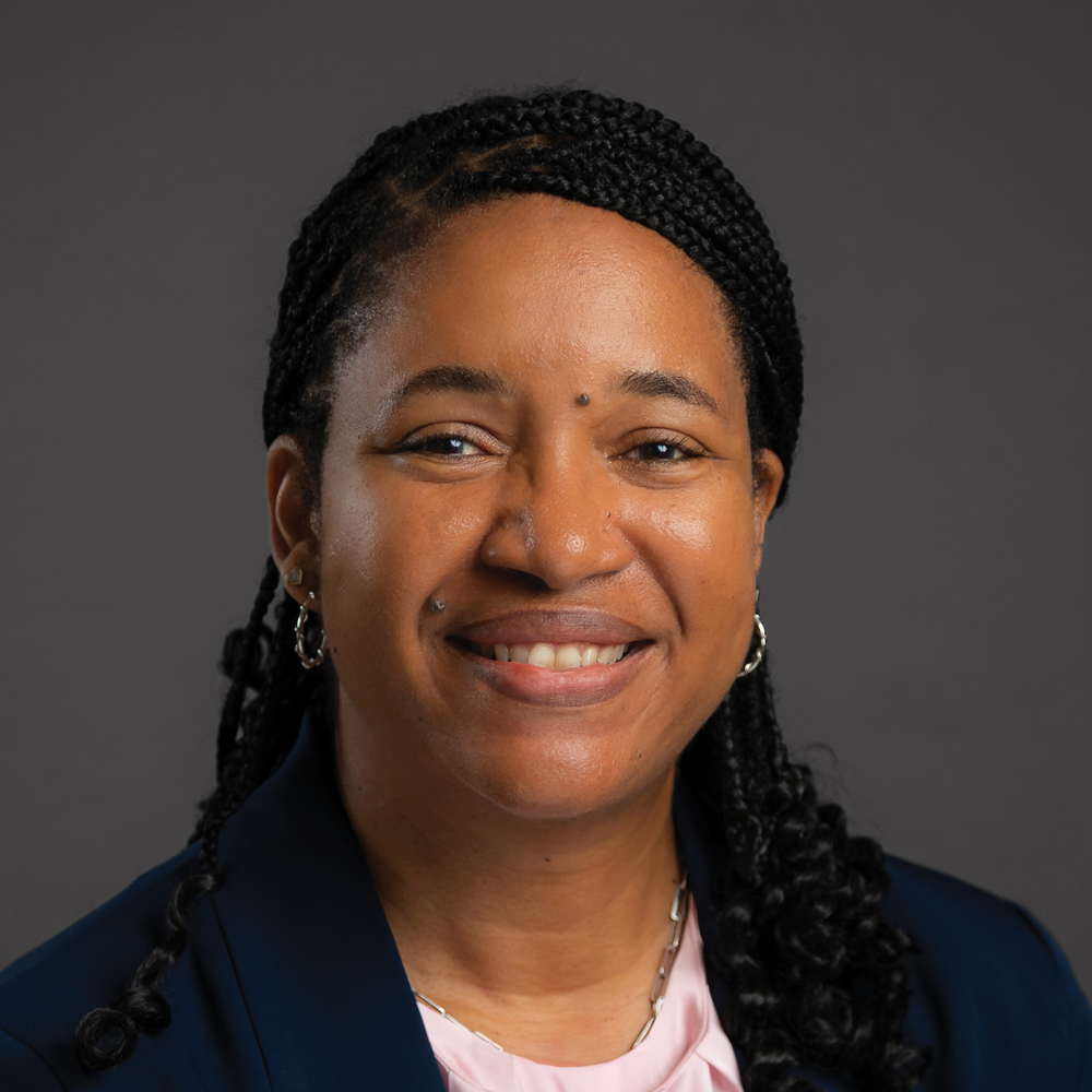 Smiling woman with long braided black hair, wearing hoop earrings, a navy blazer, and a light pink blouse against a dark gray background.