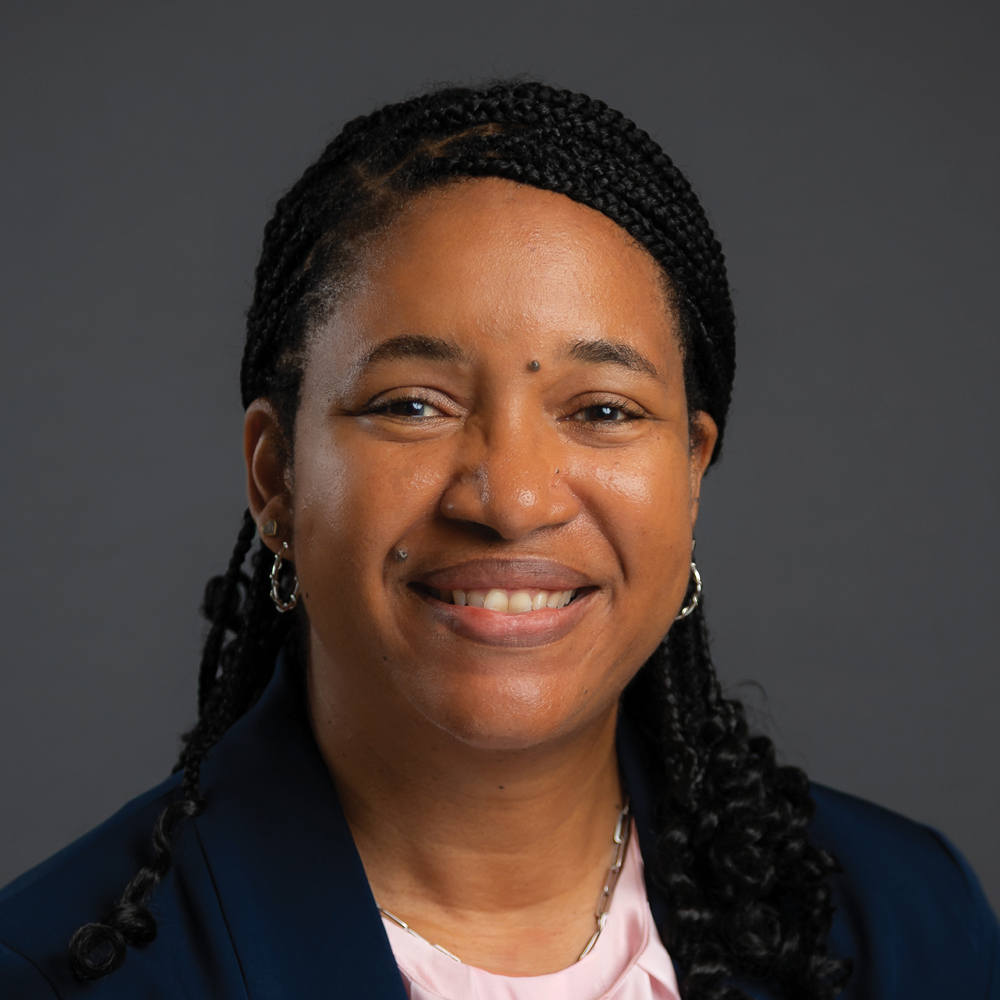 Smiling woman with long braided black hair, wearing hoop earrings, a navy blazer, and a light pink blouse against a dark gray background.