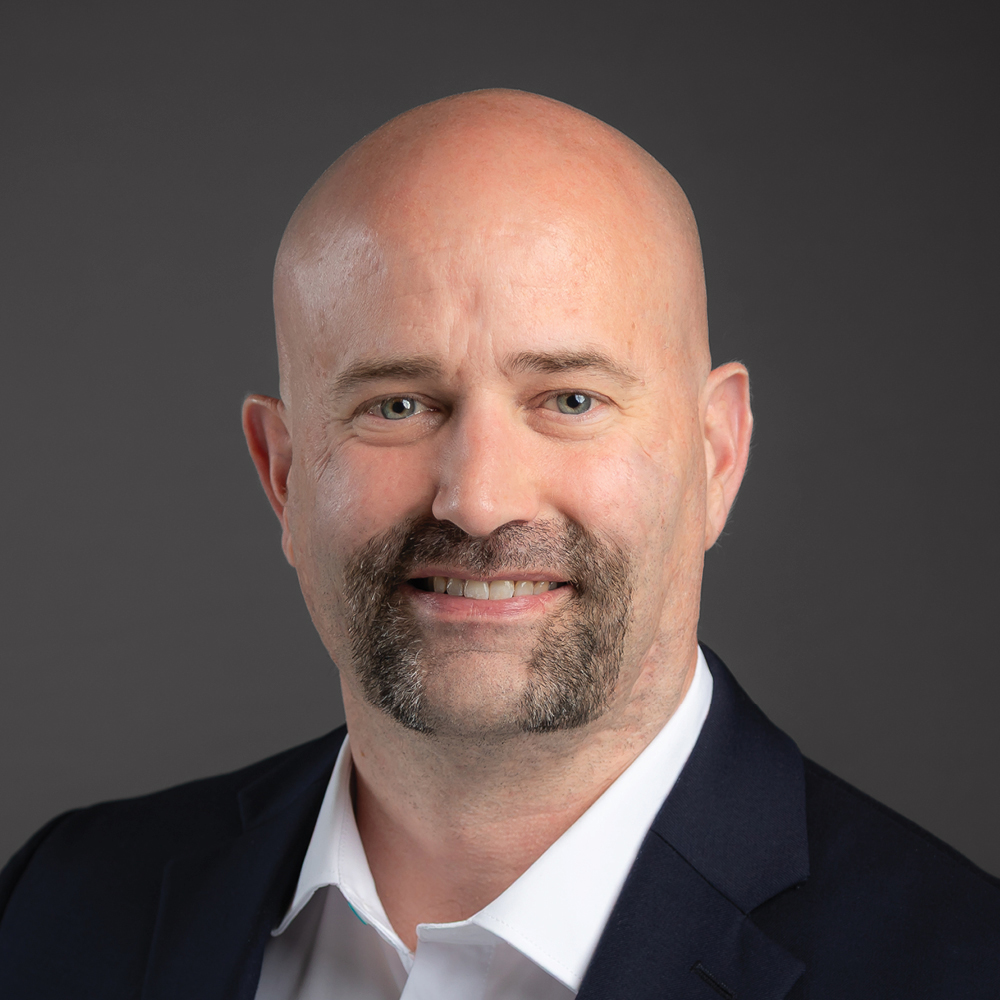 Smiling bald man with a goatee, wearing a dark blazer and white shirt against a dark gray background.
