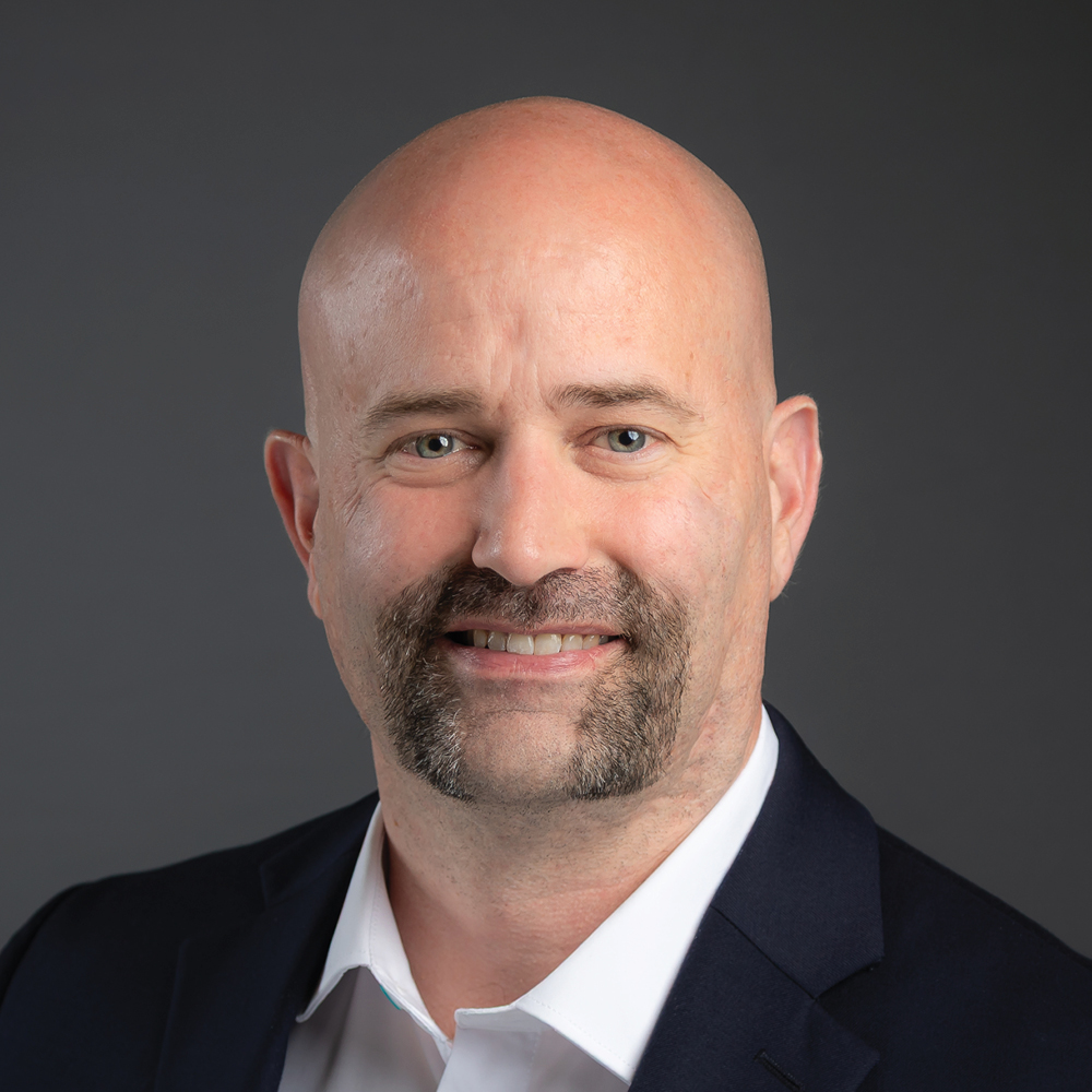 Smiling bald man with a goatee, wearing a dark blazer and white shirt against a dark gray background.