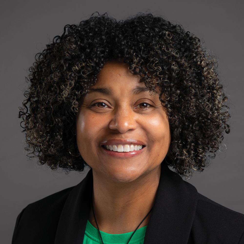 Smiling woman with short curly black hair, wearing a black blazer and green top against a dark gray background.