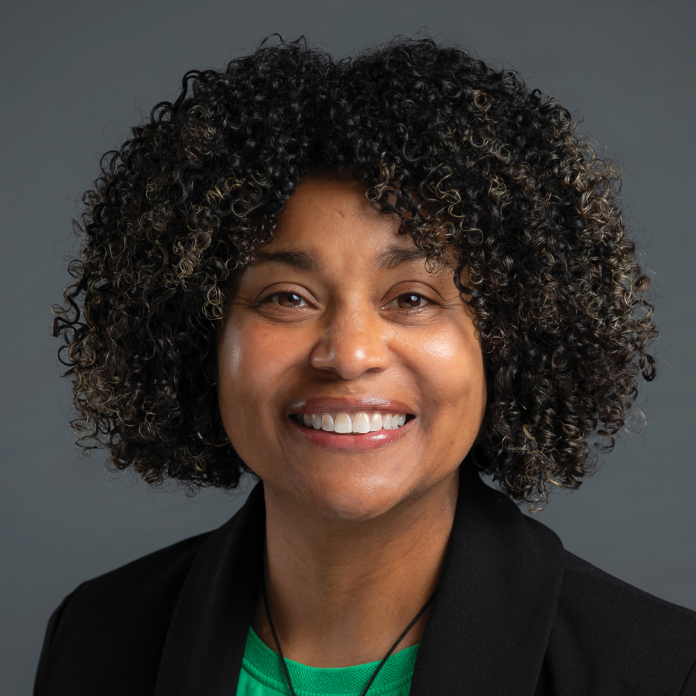 Smiling woman with short curly black hair, wearing a black blazer and green top against a dark gray background.