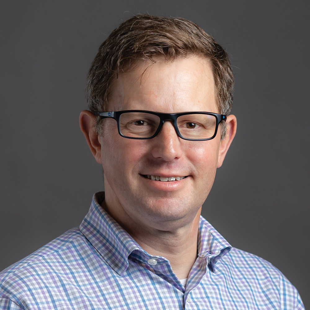 greg brubaker portrait wearing glasses and blue plaid button collar shirt