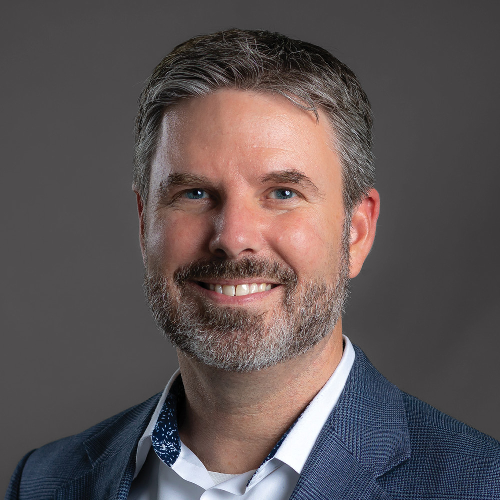 Smiling man with short salt-and-pepper hair and a neatly trimmed beard, wearing a blue blazer and white collared shirt against a dark gray background.