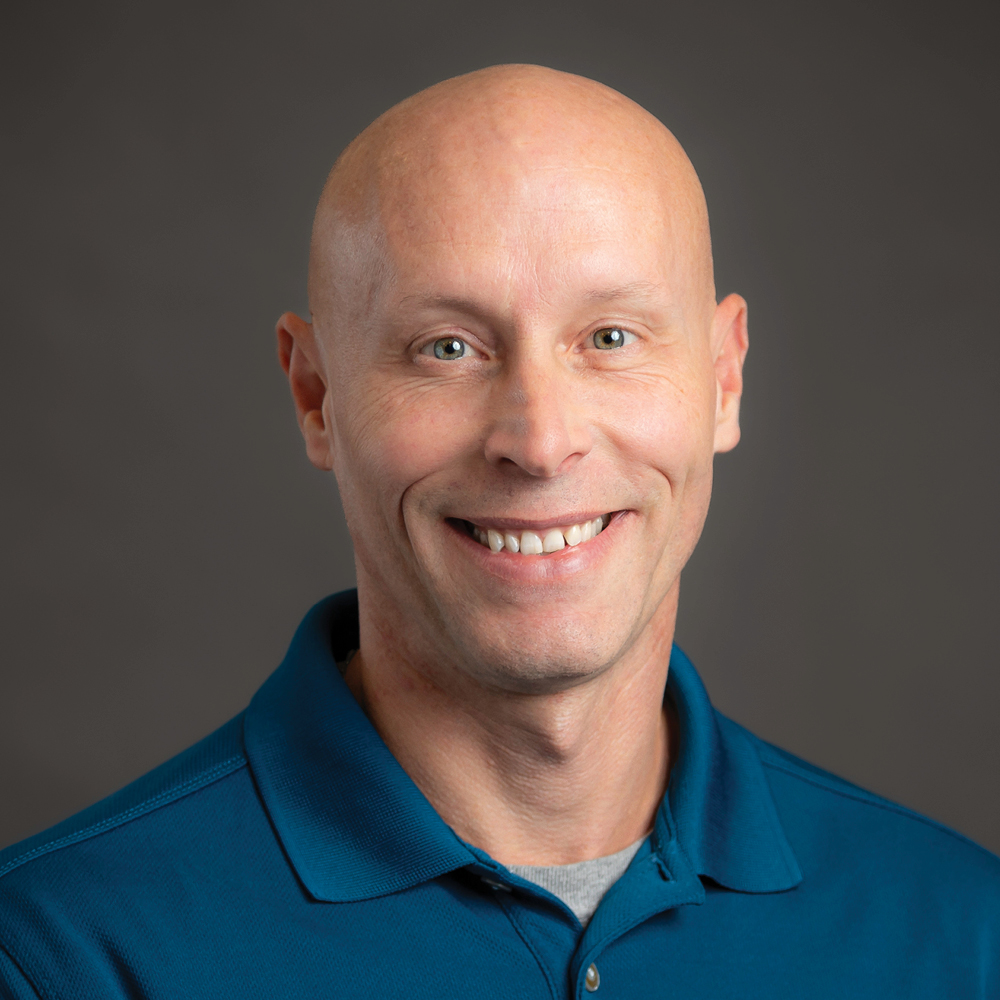 David Bolton smiling wearing blue collared shirt on dark grey background.