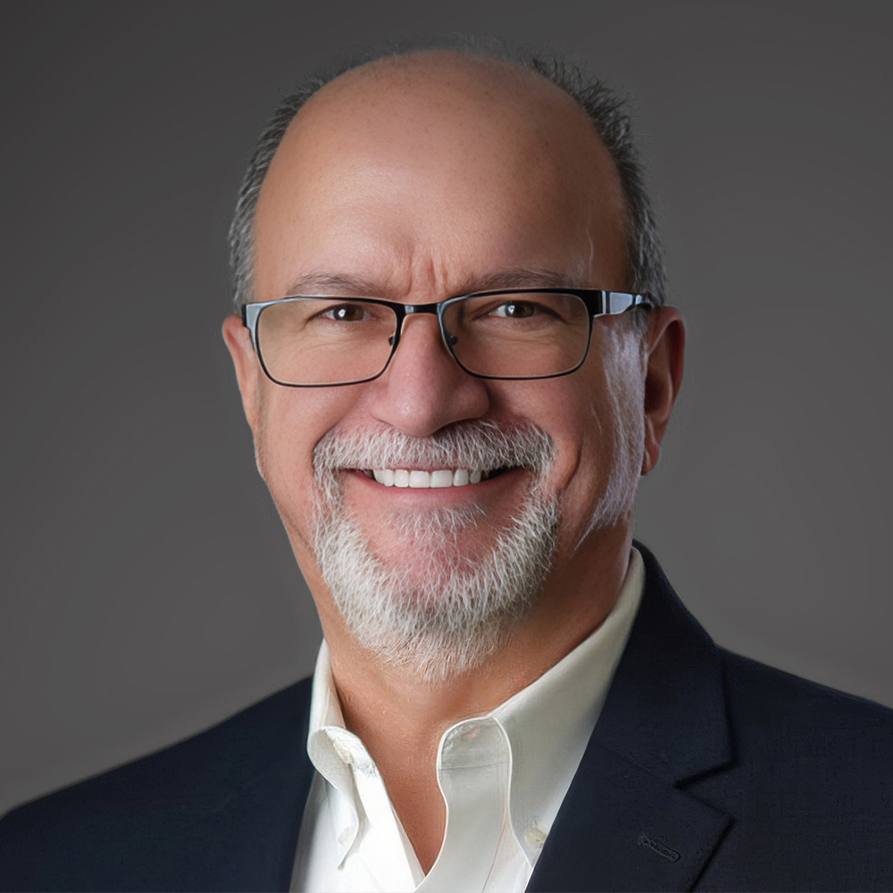 Smiling man with glasses, a white beard, and balding gray hair, wearing a dark blazer and white collared shirt against a gray background.