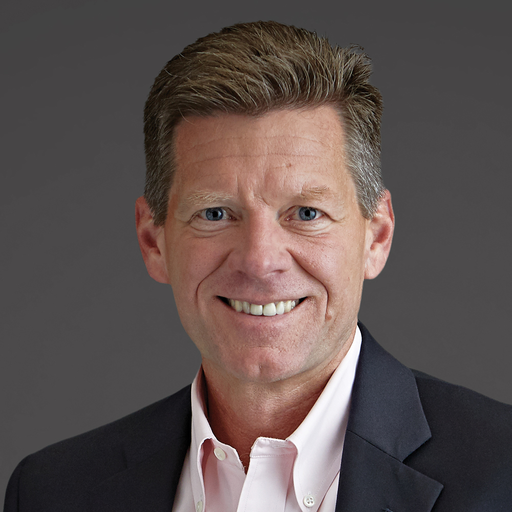 Smiling man with short brown and gray hair, wearing a dark blazer and light pink button-down shirt against a dark gray background.