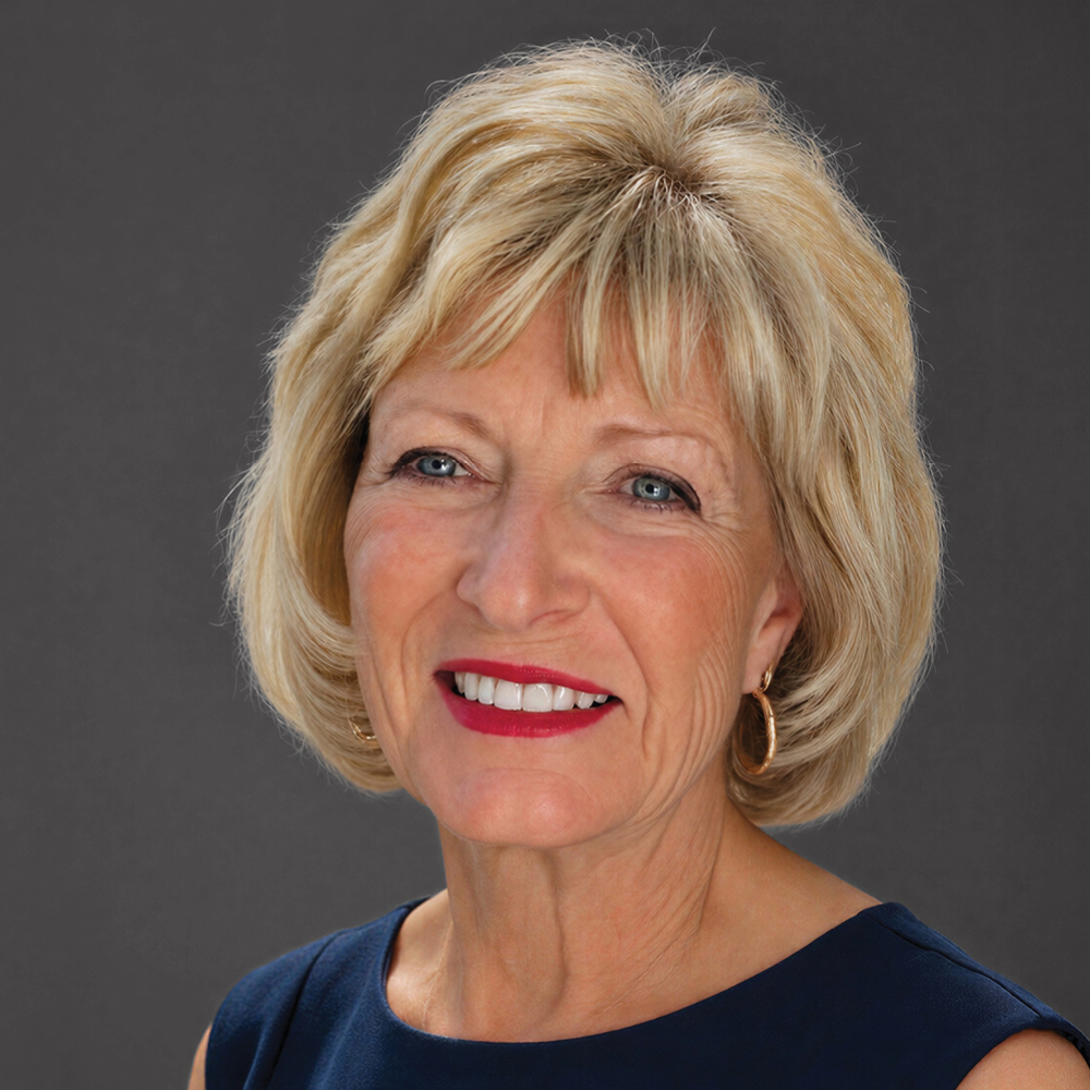 Smiling woman with short blonde hair and bangs, wearing gold hoop earrings and a navy sleeveless top against a dark gray background.