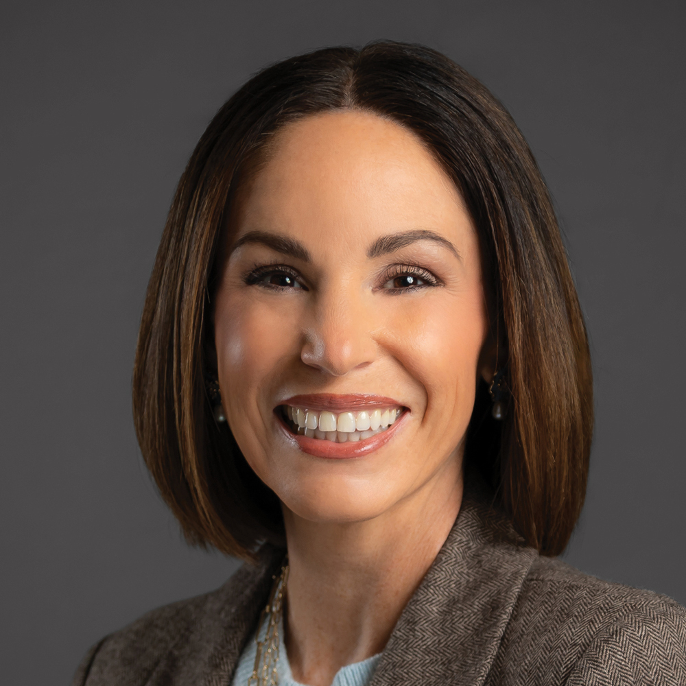 Smiling woman with straight shoulder-length brown hair, wearing a brown blazer against a dark gray background.
