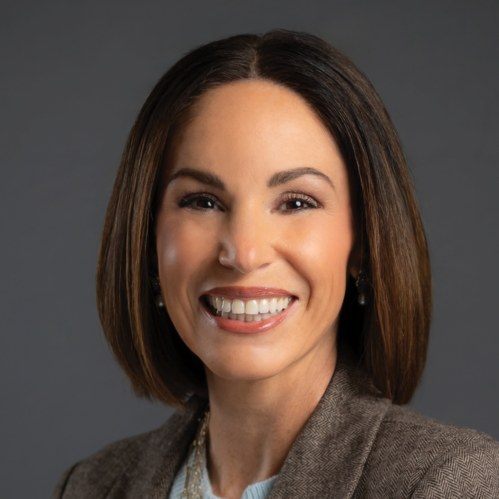 Headshot of a smiling woman with straight shoulder-length brown hair, wearing a brown blazer against a dark gray background.