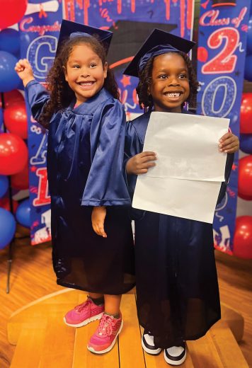 Two young children in caps and gowns celebrate their preschool graduation in front of a class graduation display.