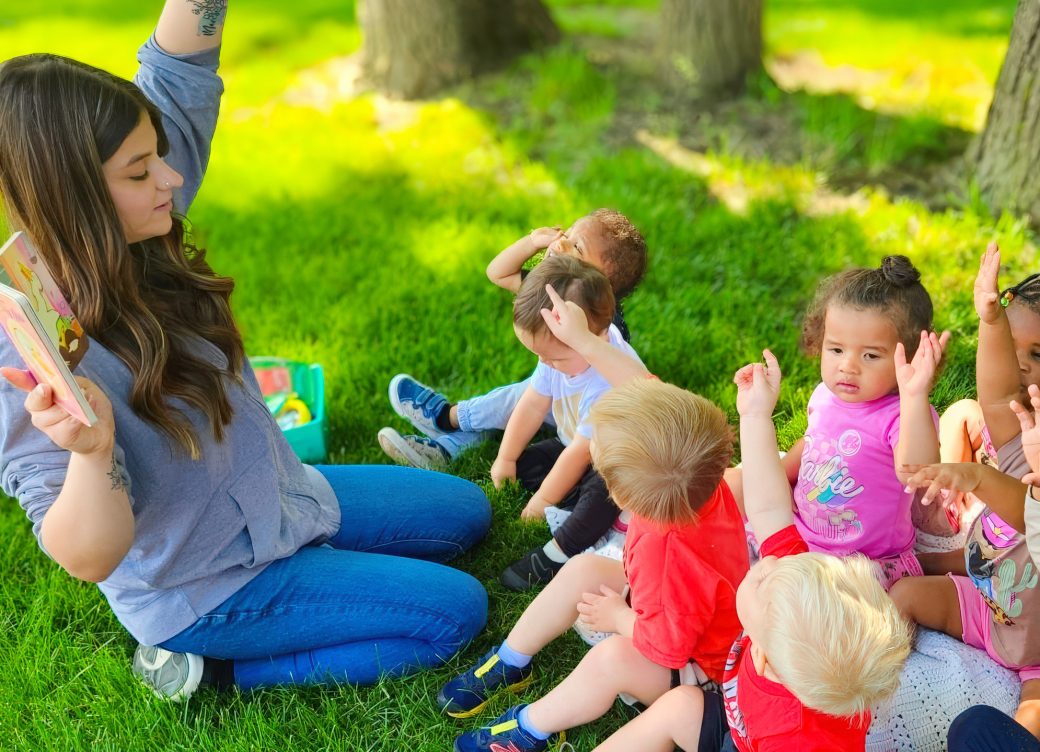 A childcare worker leads an outdoor reading activity with young children gathered on the grass.