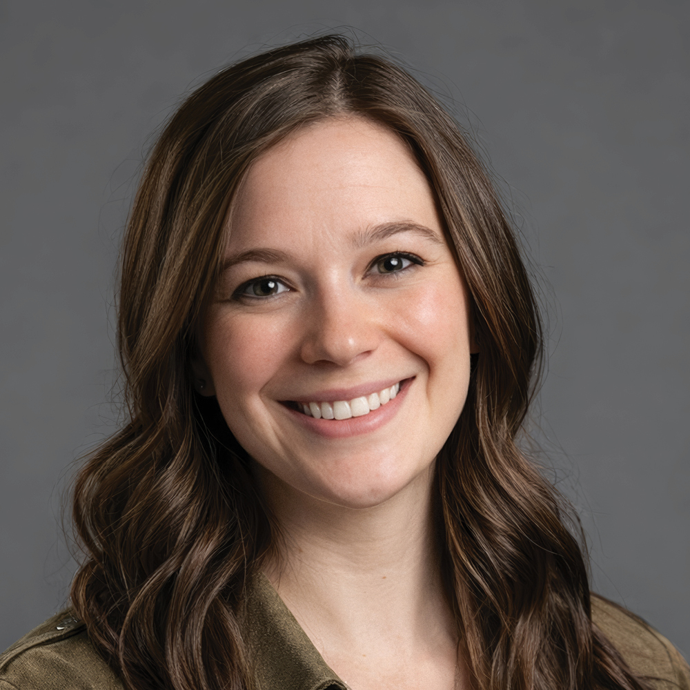 Smiling woman with long wavy brown hair, wearing an olive green collared shirt against a gray background.