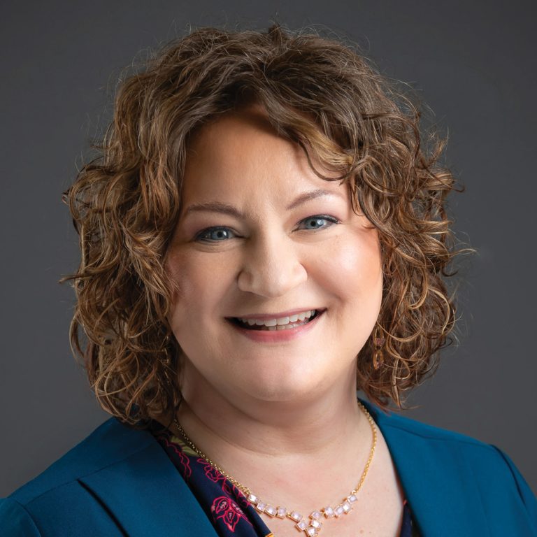 Smiling woman with curly light brown hair, wearing a teal blazer, patterned blouse, and gold necklace against a dark gray background.