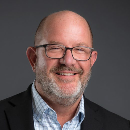 Smiling man with glasses and a gray beard, wearing a black blazer and light blue checkered shirt against a dark gray background.