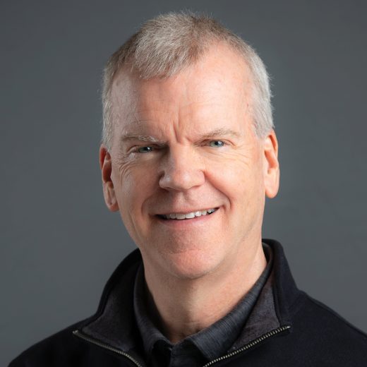 Smiling man with short gray hair, wearing a dark quarter zip-up shirt over a dark collared shirt against a dark gray background.