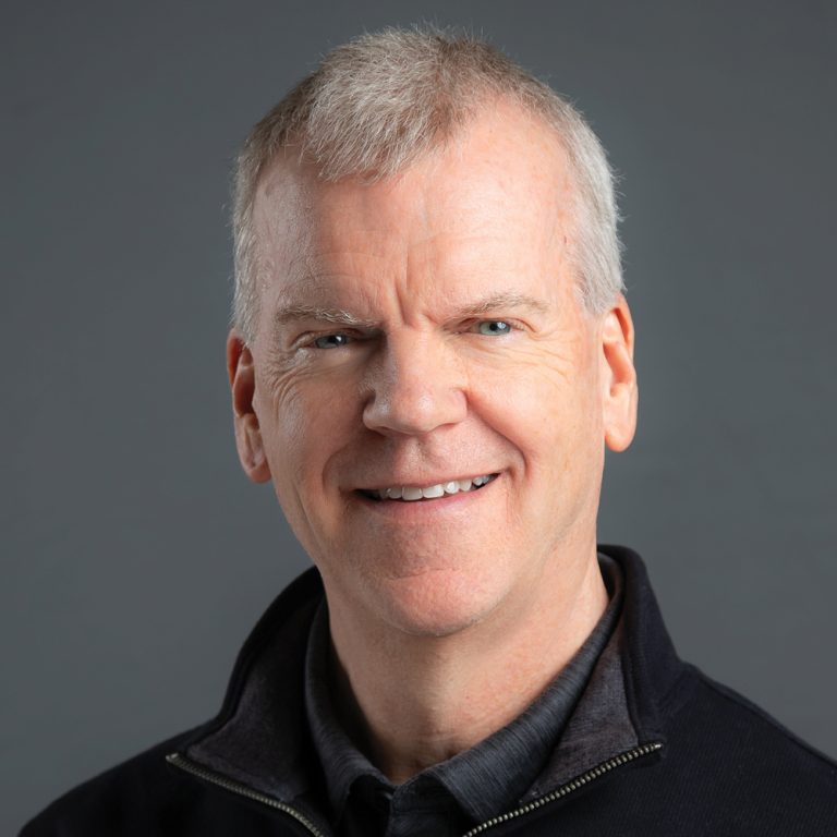 Smiling man with short gray hair, wearing a dark quarter zip-up shirt over a dark collared shirt against a dark gray background.