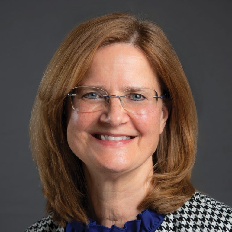 Smiling woman with shoulder-length red hair and glasses, wearing a blue blouse and black-and-white patterned jacket against a dark gray background.