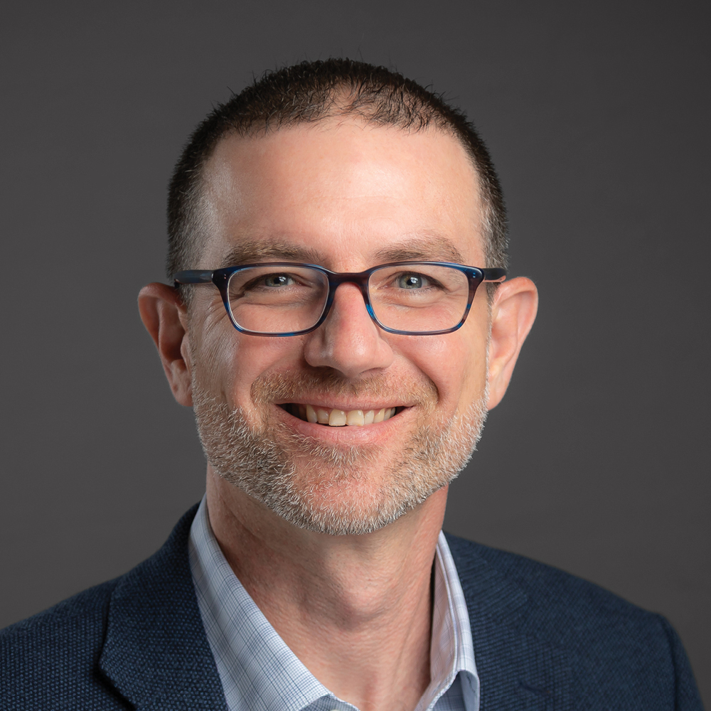 Smiling man with short dark hair, glasses, and a neatly trimmed beard, wearing a navy blazer against a dark gray background.