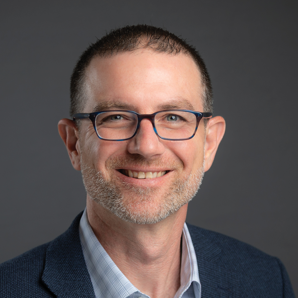 Smiling man with short dark hair, glasses, and a neatly trimmed beard, wearing a navy blazer against a dark gray background.