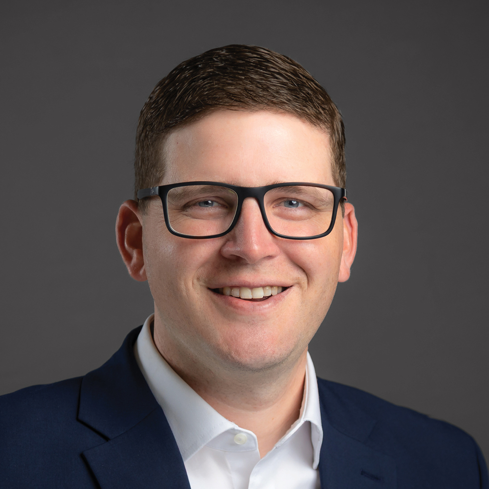 Smiling man with short brown hair and black glasses, wearing a navy blazer and white shirt against a dark gray background.