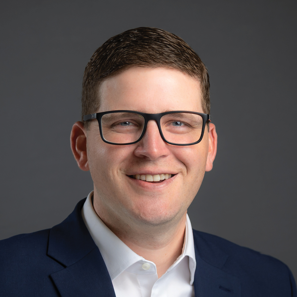 Smiling man with short brown hair and black glasses, wearing a navy blazer and white shirt against a dark gray background.