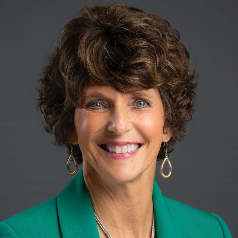 Smiling woman with short curly brown hair, wearing gold teardrop earrings and a green blazer against a dark gray background.