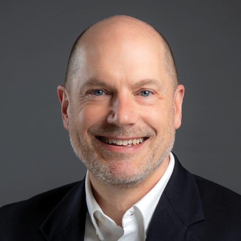 Smiling balding man with a short gray beard, wearing a dark blazer and white shirt against a dark gray background.