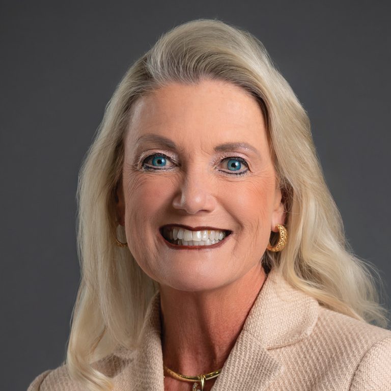 Smiling woman with long blonde hair, wearing gold jewelry and a cream blazer against a dark gray background.