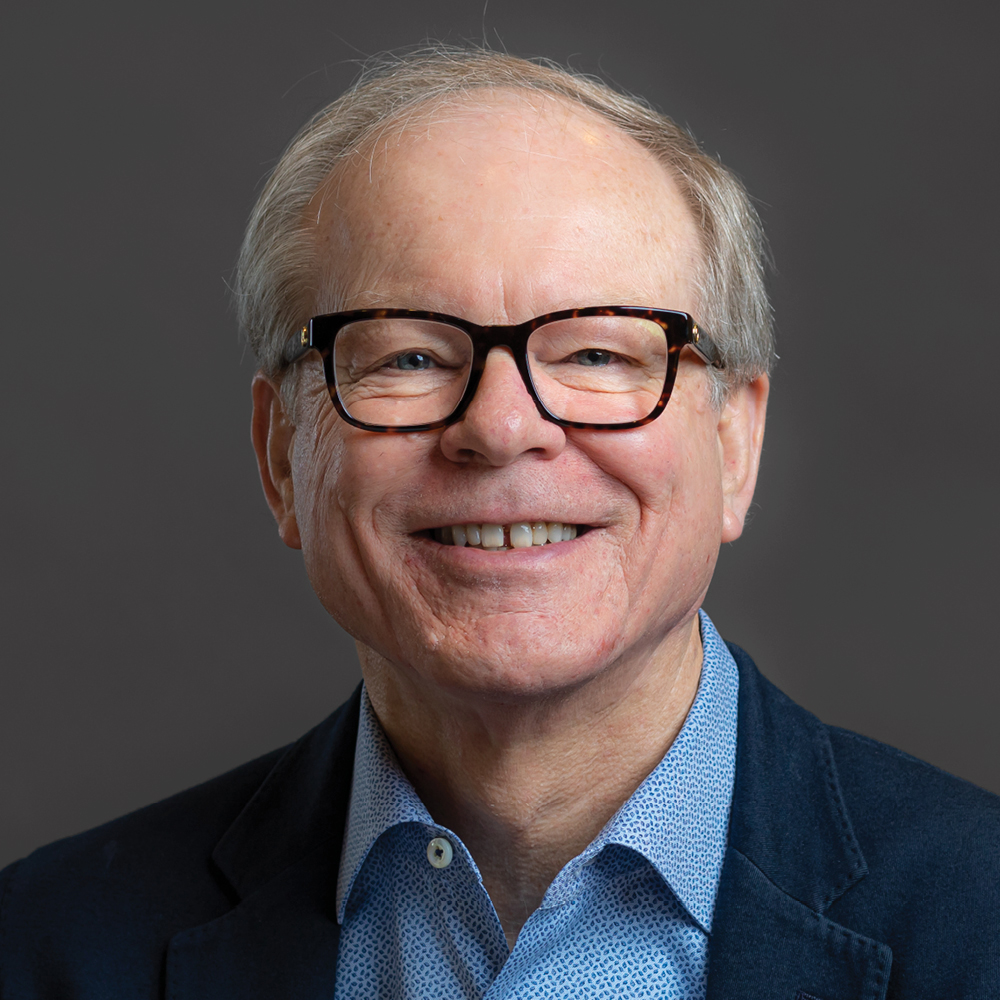 Smiling man with short gray hair and dark-framed glasses, wearing a navy blazer and light blue patterned shirt against a dark gray background.